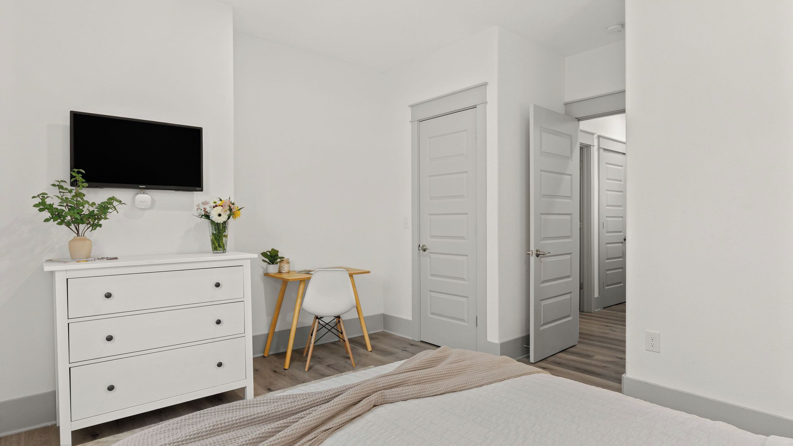 A bedroom with a bed, dresser, TV, desk, chair, and two doors.