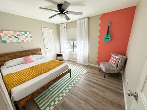 A bedroom with a bed, a ceiling fan, a window with curtains, a guitar on the wall, and a chair with a cushion.