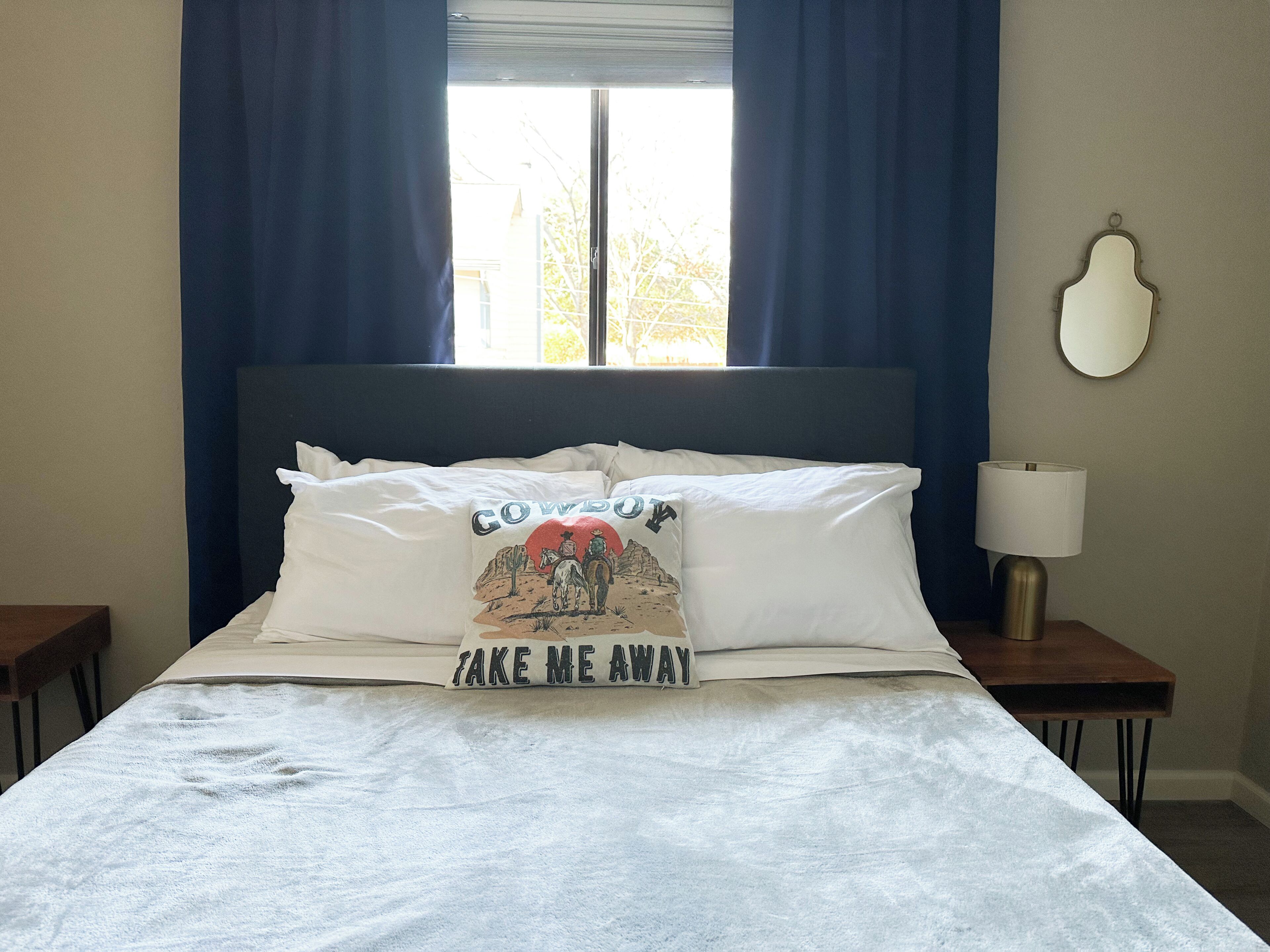 Una cama con edredón blanco, una almohada con temática vaquera y una lámpara de noche.