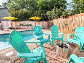 A backyard with a swimming pool, green Adirondack chairs, and a metal fire pit.