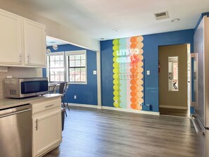 A kitchen with white cabinets, a microwave, and a window. There is a neon sign on the wall and a door leading to another room.