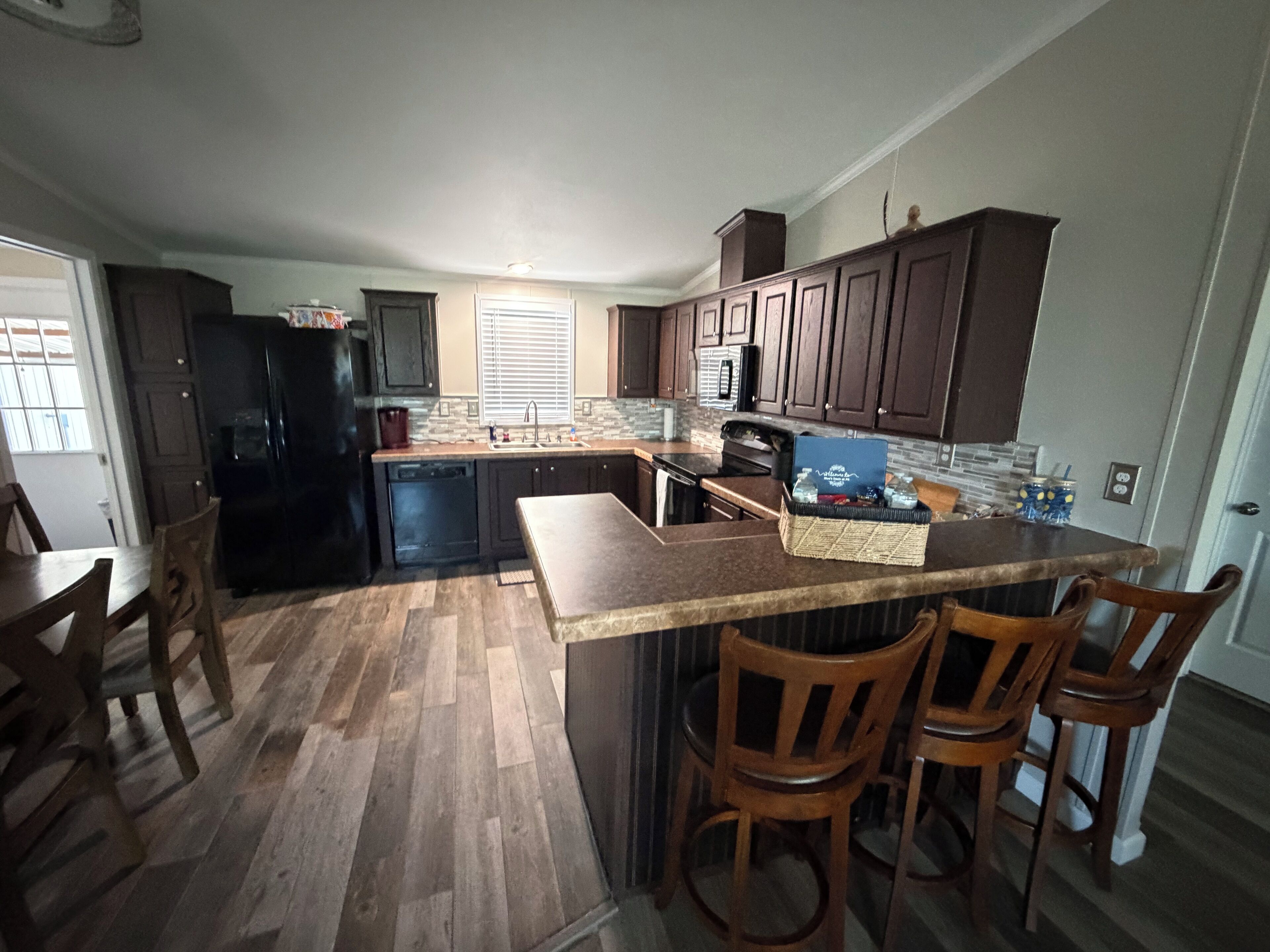 A kitchen with dark wooden cabinets, a black refrigerator, a countertop with a basket, and a dining area with wooden chairs.