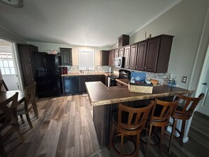 A kitchen with dark wooden cabinets, a black refrigerator, a countertop with a basket, and a dining area with wooden chairs. - Blue’s Oasis- less than 15 minutes from Rocker B Ranch (Graford)