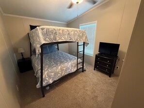 A bunk bed with a patterned bedspread, a bedside table with a lamp, a dresser with a television, and a window with blinds. - Blue’s Oasis- less than 15 minutes from Rocker B Ranch (Graford)