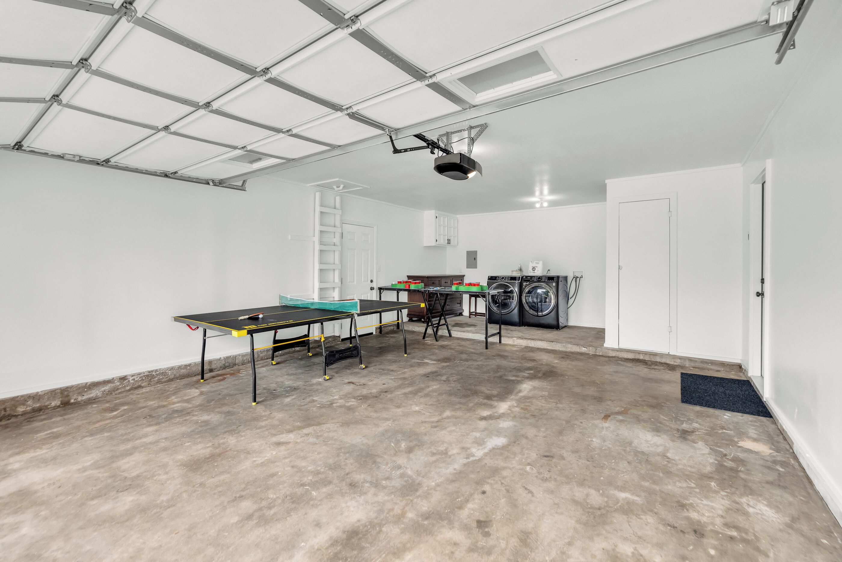 Amplio espacio interior con mesa de ping pong, lavadora y una escalera apoyada en una pared blanca.
