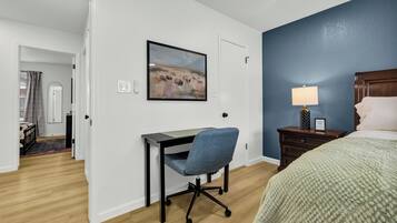 A hotel room with a desk, chair, bed, nightstand, lamp, and a painting on the wall.