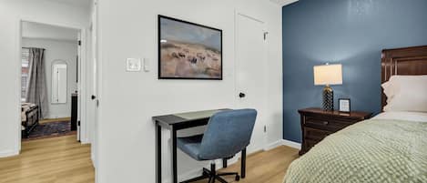 Habitación de hotel con escritorio, silla, cama, mesita de noche, lámpara y un cuadro en la pared.