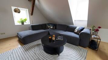 Ein modernes Wohnzimmer mit einem Ecksofa, einem runden Couchtisch, einem kleinen Beistelltisch mit einer Vase und einem Fenster mit Pflanzen.