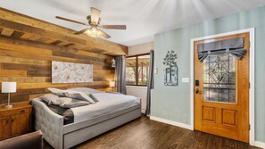 A bedroom with a bed, wooden wall paneling, a ceiling fan, a door with a window, and a wall-mounted light fixture.