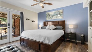 A bedroom with a large bed, a ceiling fan, a nightstand with a lamp, and a painting on the wall.