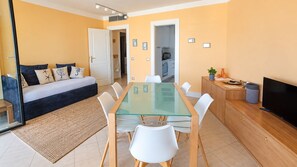 Sala de estar moderna com mesa de jantar de vidro, cadeiras brancas, sofá com almofadas decorativas, televisão e área de cozinha visível através de uma porta aberta.