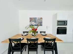 A dining area with a wooden table, black chairs, and a centerpiece with flowers. There is a painting on the wall and a kitchen with a microwave and oven. - Modern/Lux penthouse In front of rooftop pool (Las Terrenas)