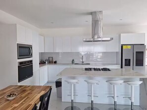 A modern kitchen with white cabinetry, a built-in oven, a stainless steel range hood, a double sink, and a central island with bar stools. - Modern/Lux penthouse In front of rooftop pool (Las Terrenas)
