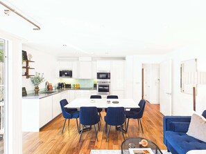 A modern kitchen with white cabinets, a dining table with chairs, and a blue sofa in the living area. - Westminster Spacious 2BR Apartment with Balcony (Greater London)