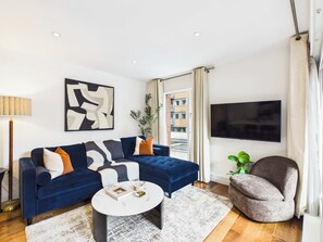 A modern living room with a blue sofa, a round coffee table, a grey armchair, a flat-screen TV, and a large abstract painting. - Westminster Spacious 2BR Apartment with Balcony (Greater London)