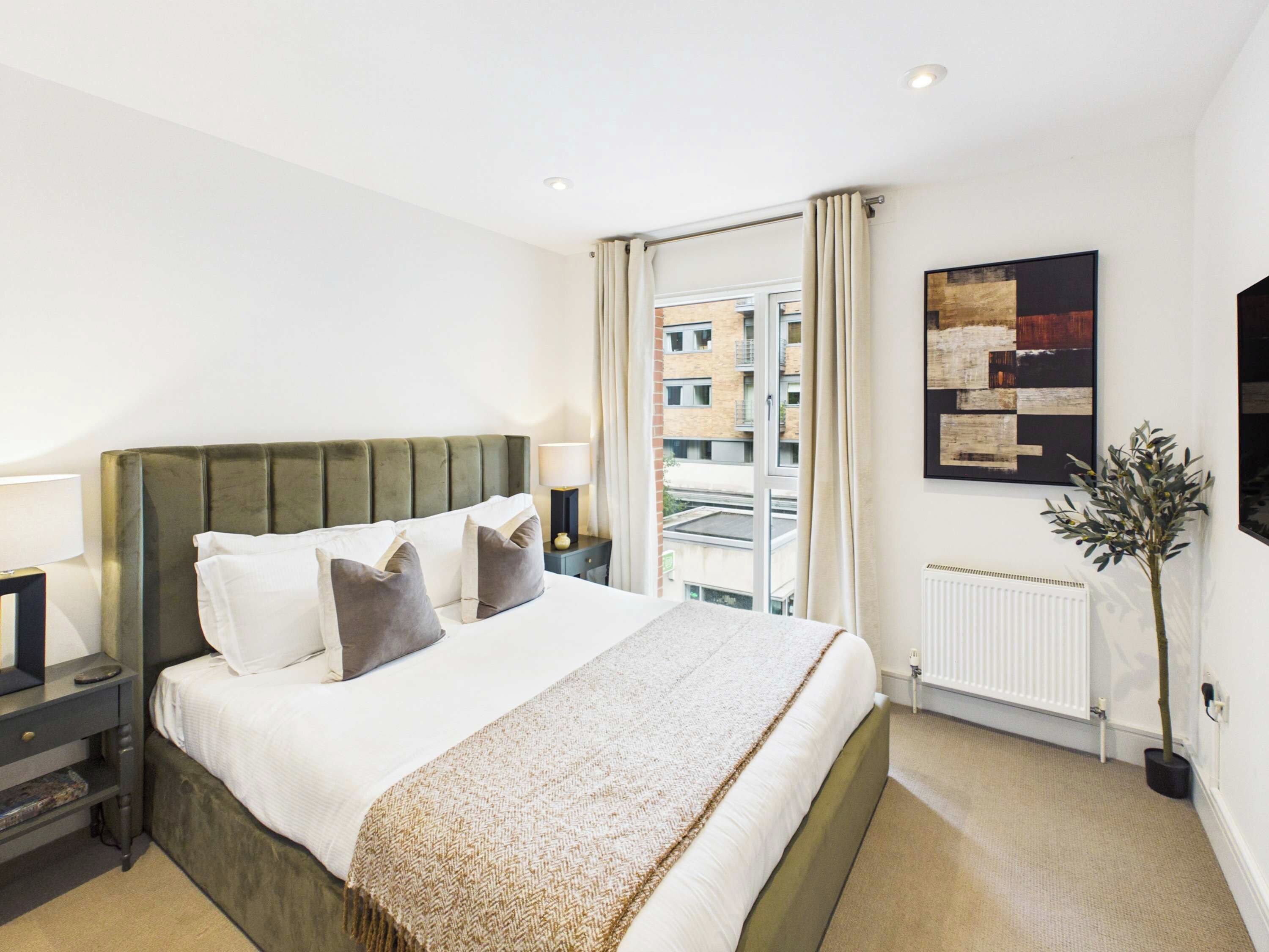 A hotel room with a large bed, a nightstand, a lamp, a window with curtains, a radiator, and a wall-mounted artwork.