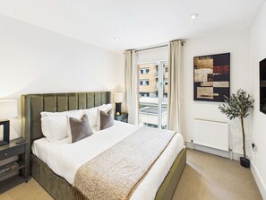 Een hotelkamer met een groot bed, een nachtkastje, een lamp, een raam met gordijnen, een radiator en een aan de muur hangend kunstwerk.