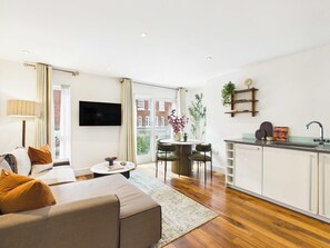 A modern living room with a sofa, a coffee table, and a TV. There is a dining area with a table and chairs, and a kitchen with a sink and cabinets. - Bright One Bedroom Apartment in Westminster (Greater London)