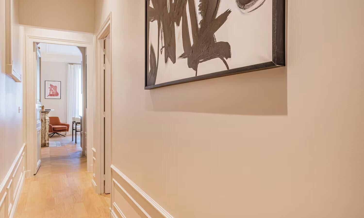A hallway with a framed abstract painting, a door leading to another room, and a chair in the distance.