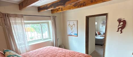 A bedroom with a bed, a window with curtains, a wooden ceiling beam, and a door leading to another room.