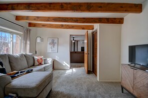 A living room with a grey sofa, a television on a wooden stand, and a wooden ceiling beam. - Authentic Adobe Casita near Old Town - King Bed (Albuquerque)