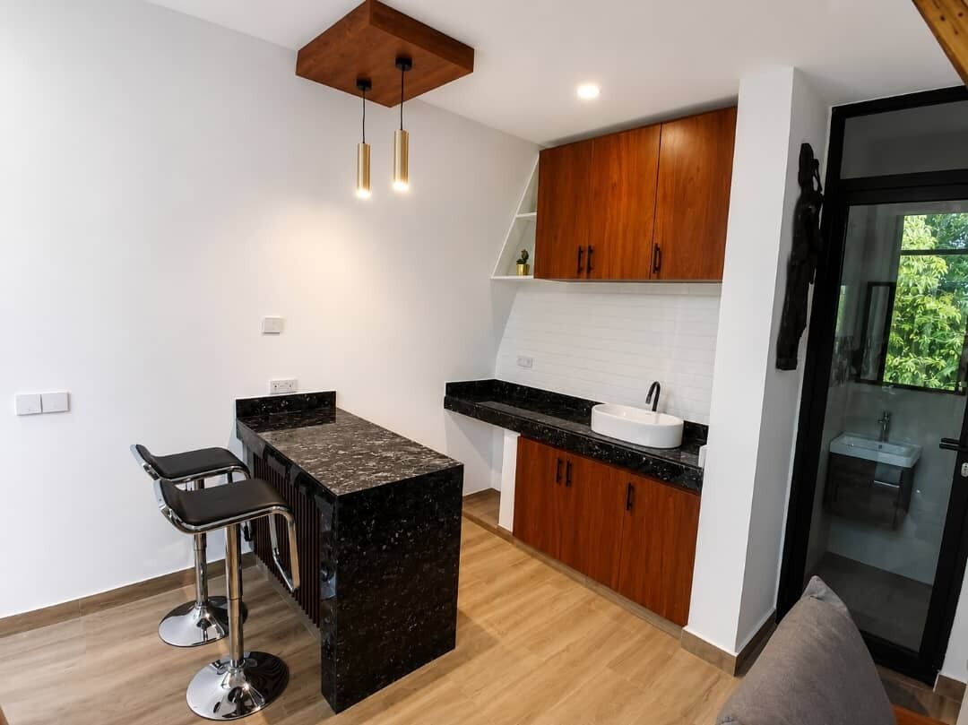 A modern kitchen with a central island, bar stools, wooden cabinets, and a glass-enclosed bathroom.