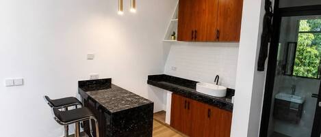 A modern kitchen with a central island, bar stools, wooden cabinets, and a glass-enclosed bathroom.