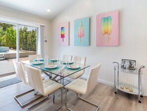 A dining area with a glass table, white chairs, and a glass cart with glasses and a picture frame. - Modern Palm Springs Retreat with Heated Pool (Palm Springs)