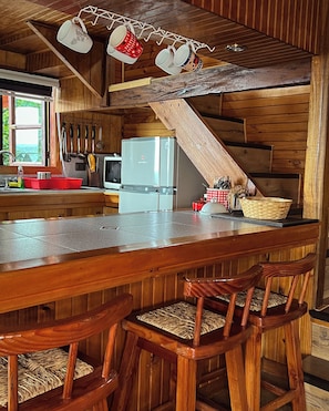 A rustic kitchen with wooden walls, a bar area with stools, and hanging mugs. - Cabañas Ruta Del Lago (LOS RISCOS)