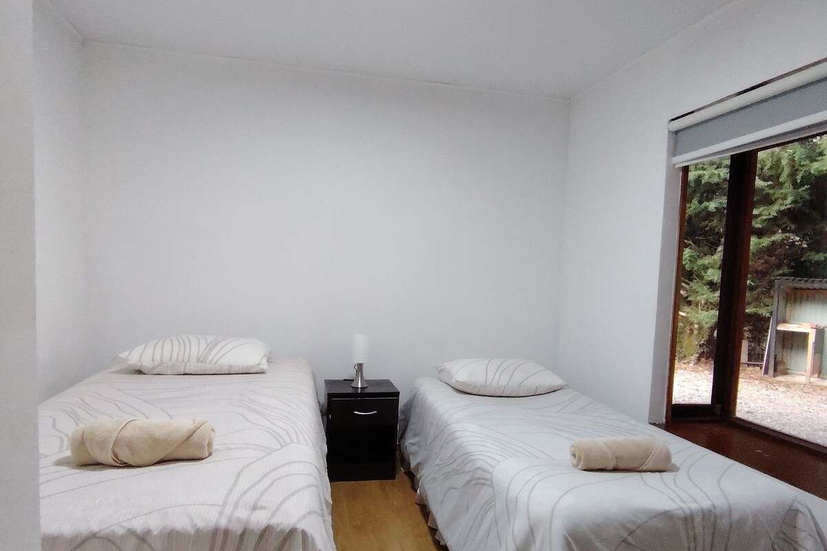 A room with two single beds, each with a pillow and a rolled towel, a bedside table with a lamp, and a window with a view of greenery.