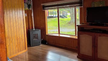 Interior de una cabaña con estufa de leña, televisor y un ventanal con vistas a un área verde.