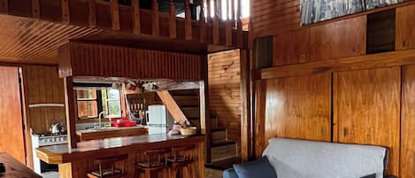 A cozy cabin-style interior with a wooden ceiling, a kitchen area with a stove and sink, a bar with stools, and a blue sofa.