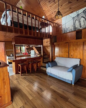 A cozy cabin-style interior with a wooden ceiling, a kitchen area with a stove and sink, a bar with stools, and a blue sofa.