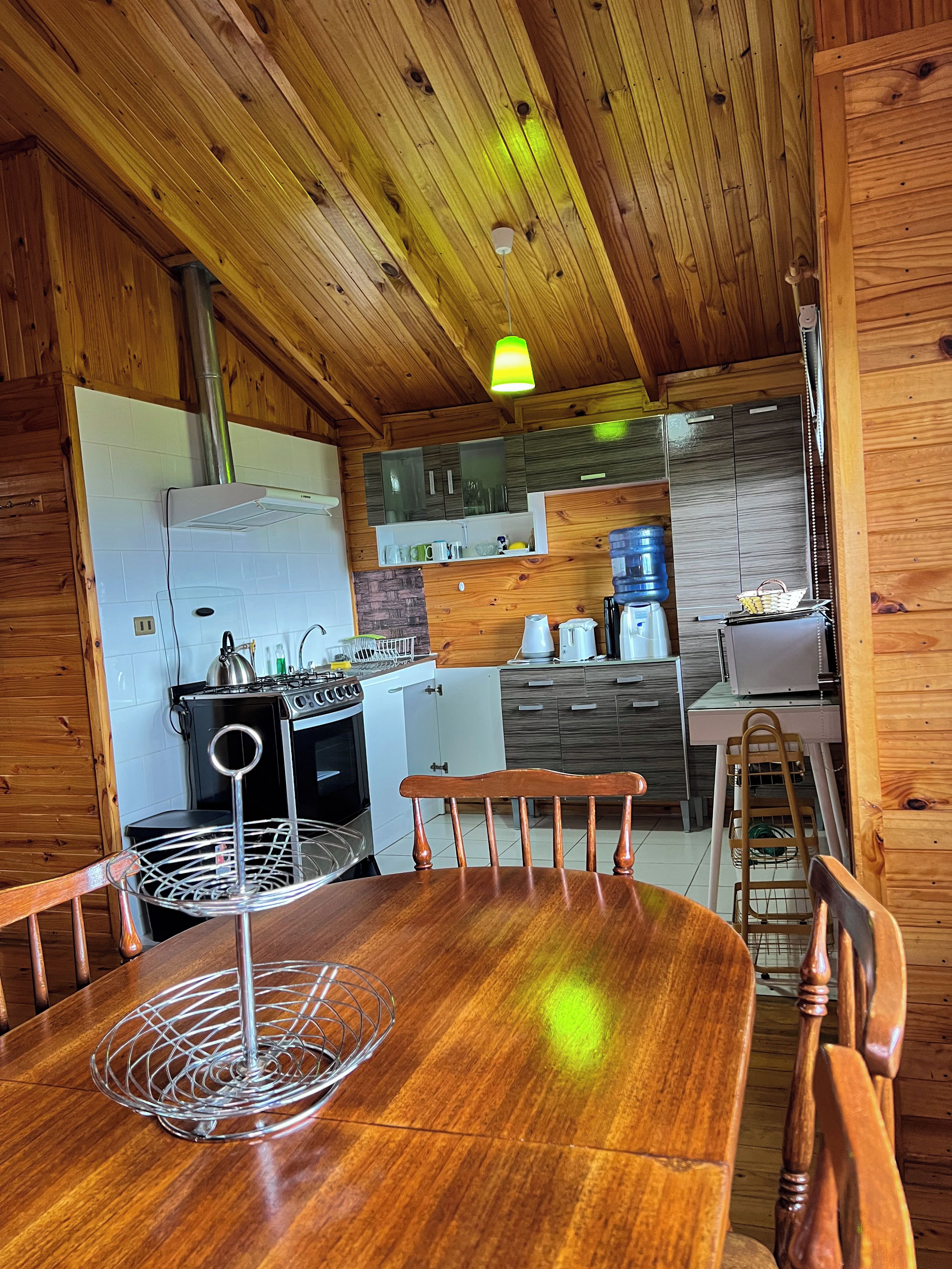 A wooden interior with a dining table, chairs, a kitchen area with appliances, and a hanging light fixture.