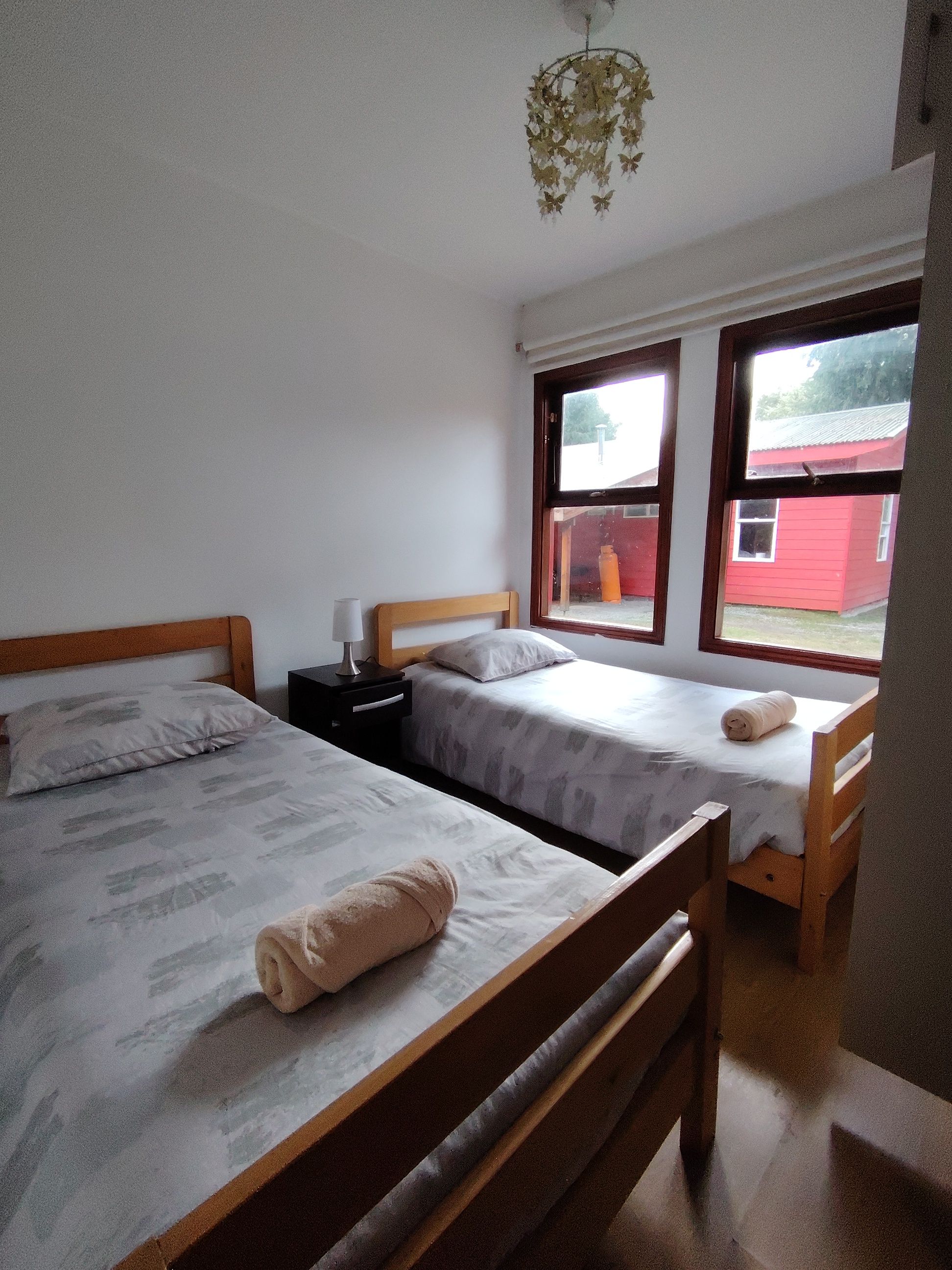 A room with two single beds, a bedside table with a lamp, a window with a view of a red building, and a hanging light fixture.