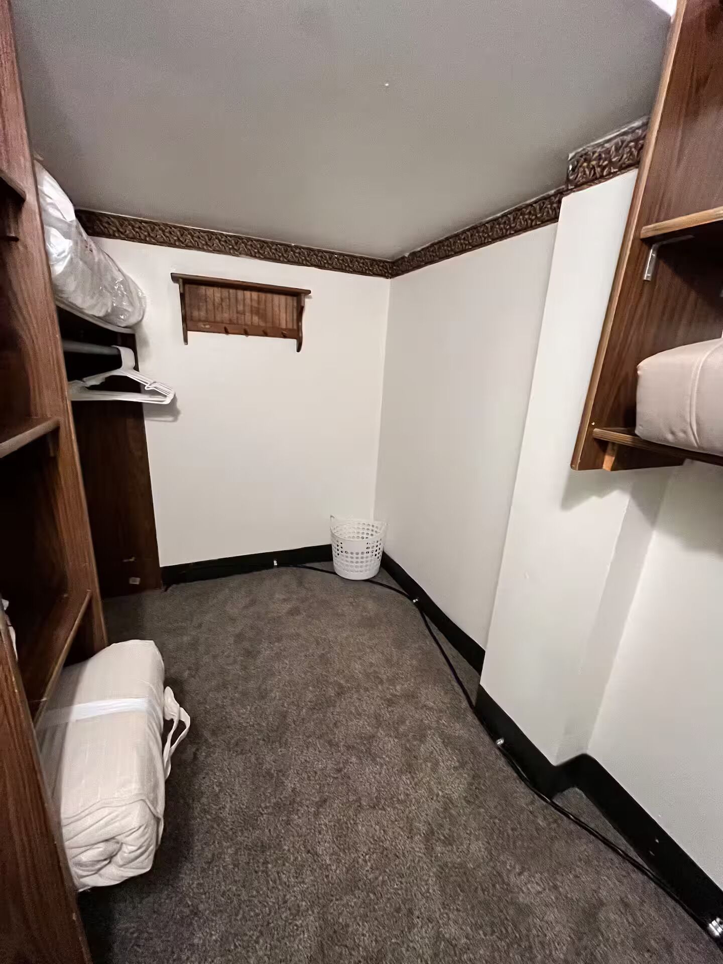 A small, carpeted room with a wooden wardrobe, a white laundry basket, and a small window.