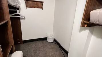 A small, carpeted room with a wooden wardrobe, a white laundry basket, and a small window.
