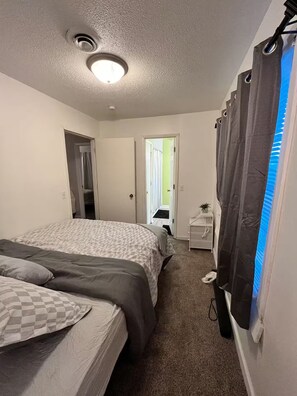 Ein Schlafzimmer mit zwei Betten, einer Deckenleuchte, einer Tür zu einem anderen Raum und einem Fenster mit Vorhängen.