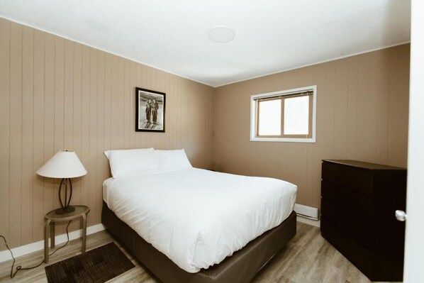 A bedroom with a bed, a bedside table with a lamp, a dresser, a window, and a wall-mounted picture.
