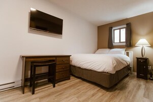 Un dormitorio con cama, escritorio, silla, televisor montado en la pared, ventana con cortinas y lámpara de noche.