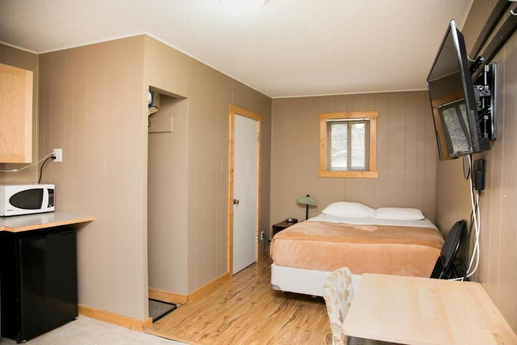A compact hotel room with a bed, a desk with a microwave, a TV mounted on the wall, and a window with blinds.