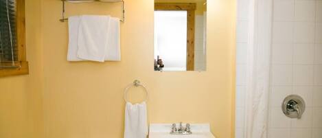 Ein Badezimmer mit Toilette, Waschbecken und Dusche. Handtücher liegen auf einem Wäscheständer, und über dem Waschbecken hängt ein Spiegel.