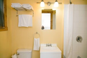 Un baño con inodoro, lavamanos y ducha. Hay toallas en un perchero y un espejo sobre el lavamanos.