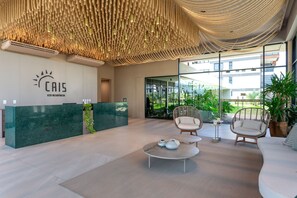 A modern reception area with a green reception desk, wicker chairs, a round coffee table, and a ceiling with a unique textured design. - Meu Xilo Hospedagem (N. Sra. do Ó)