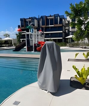 A swimming pool with a covered lounge chair, a slide, and a modern building in the background. - Meu Xilo Hospedagem (N. Sra. do Ó)