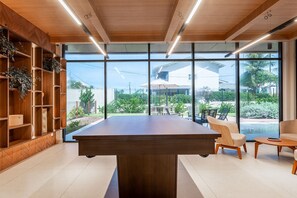A modern interior with a central wooden table, built-in shelving with plants, and large windows offering a view of a residential area. - Meu Xilo Hospedagem (N. Sra. do Ó)