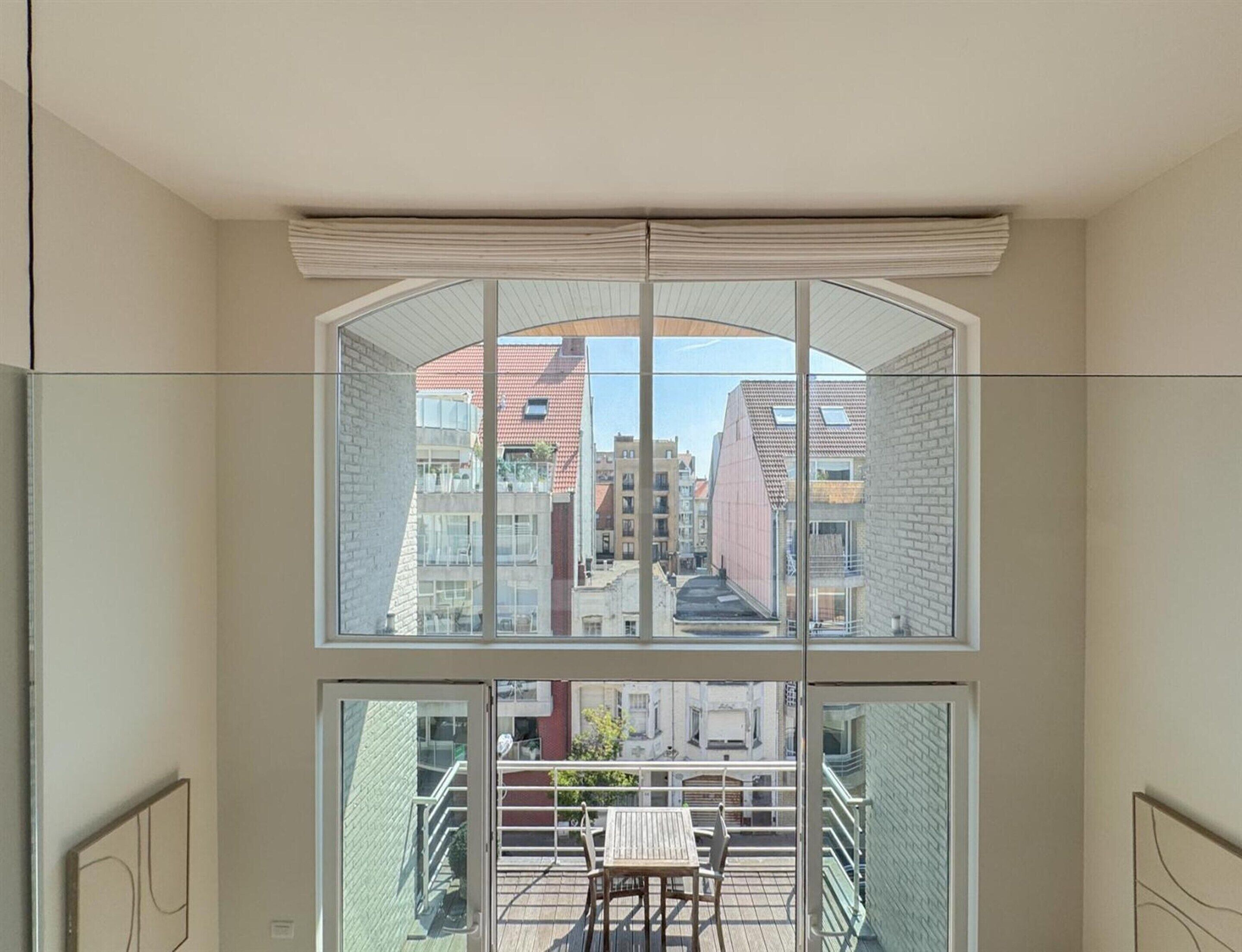 A room with a large window overlooking a cityscape, a balcony with a table and chairs, and a modern interior design.