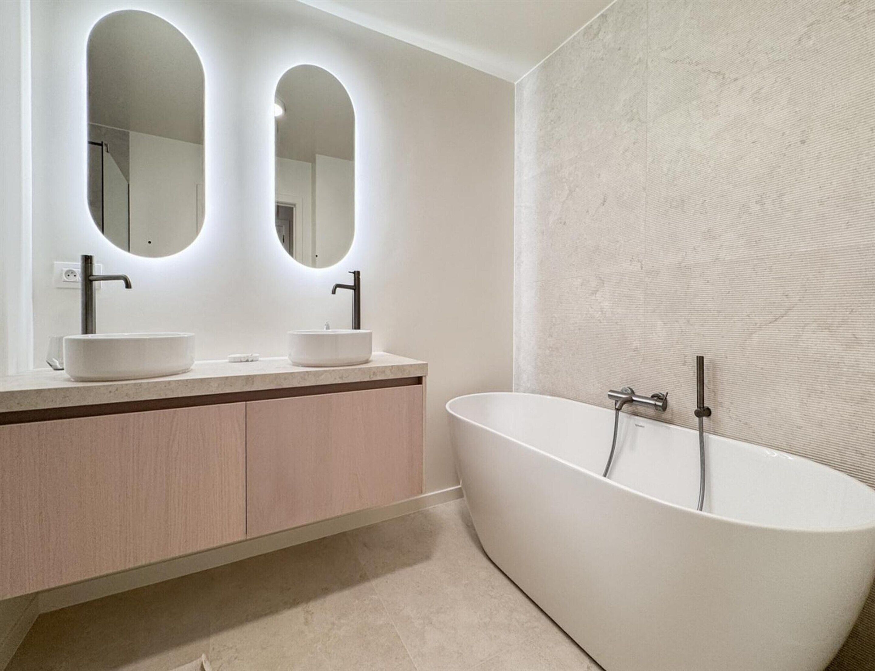 A modern bathroom with a freestanding bathtub, two sinks with round mirrors, and a textured wall.