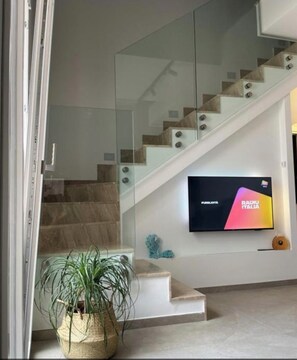Un interior moderno con una escalera, un televisor de pantalla plana mostrando RADIO ITALIA y una planta en maceta.