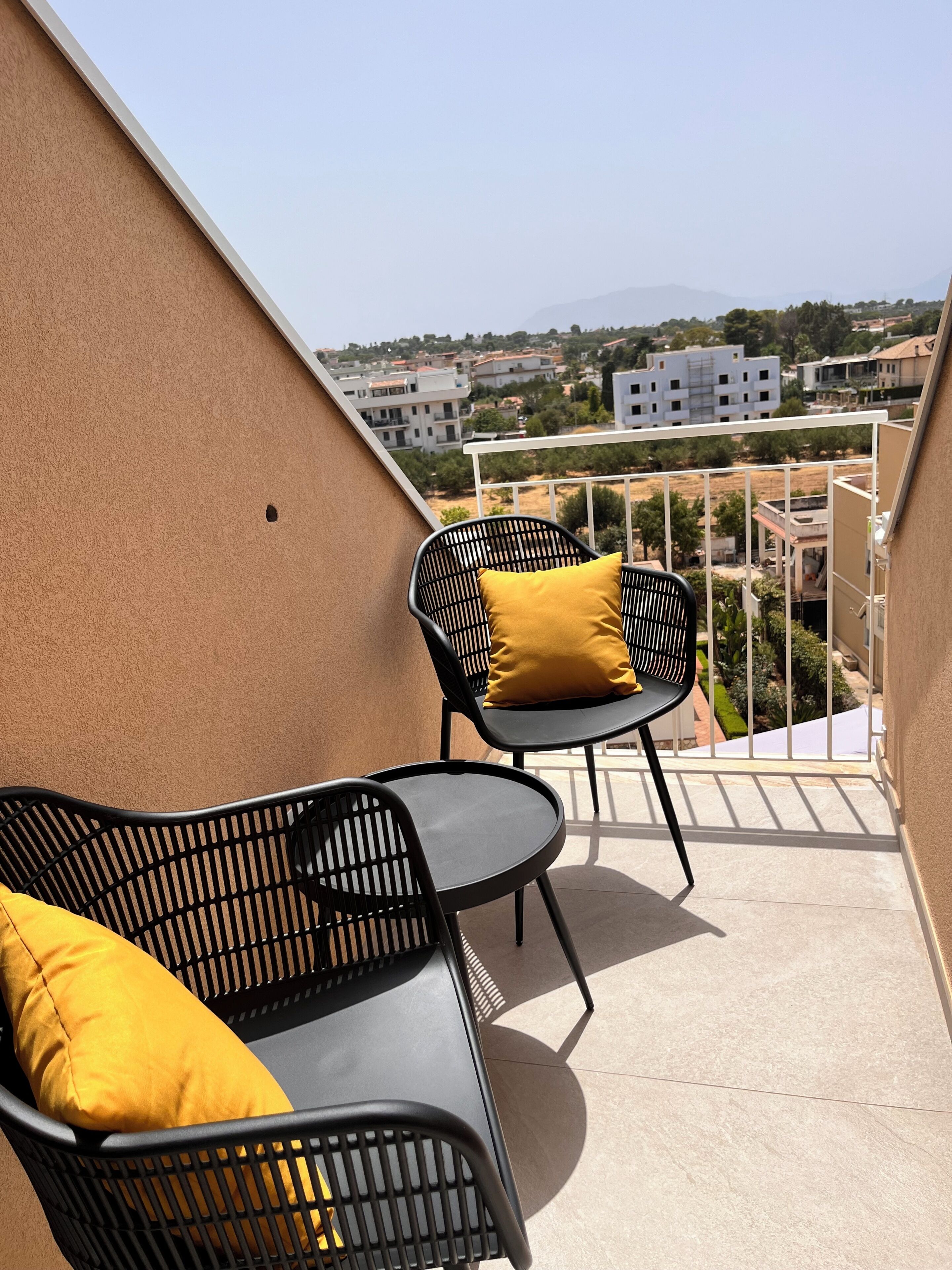 A balcony with two black metal chairs and a small table, one with a yellow cushion.